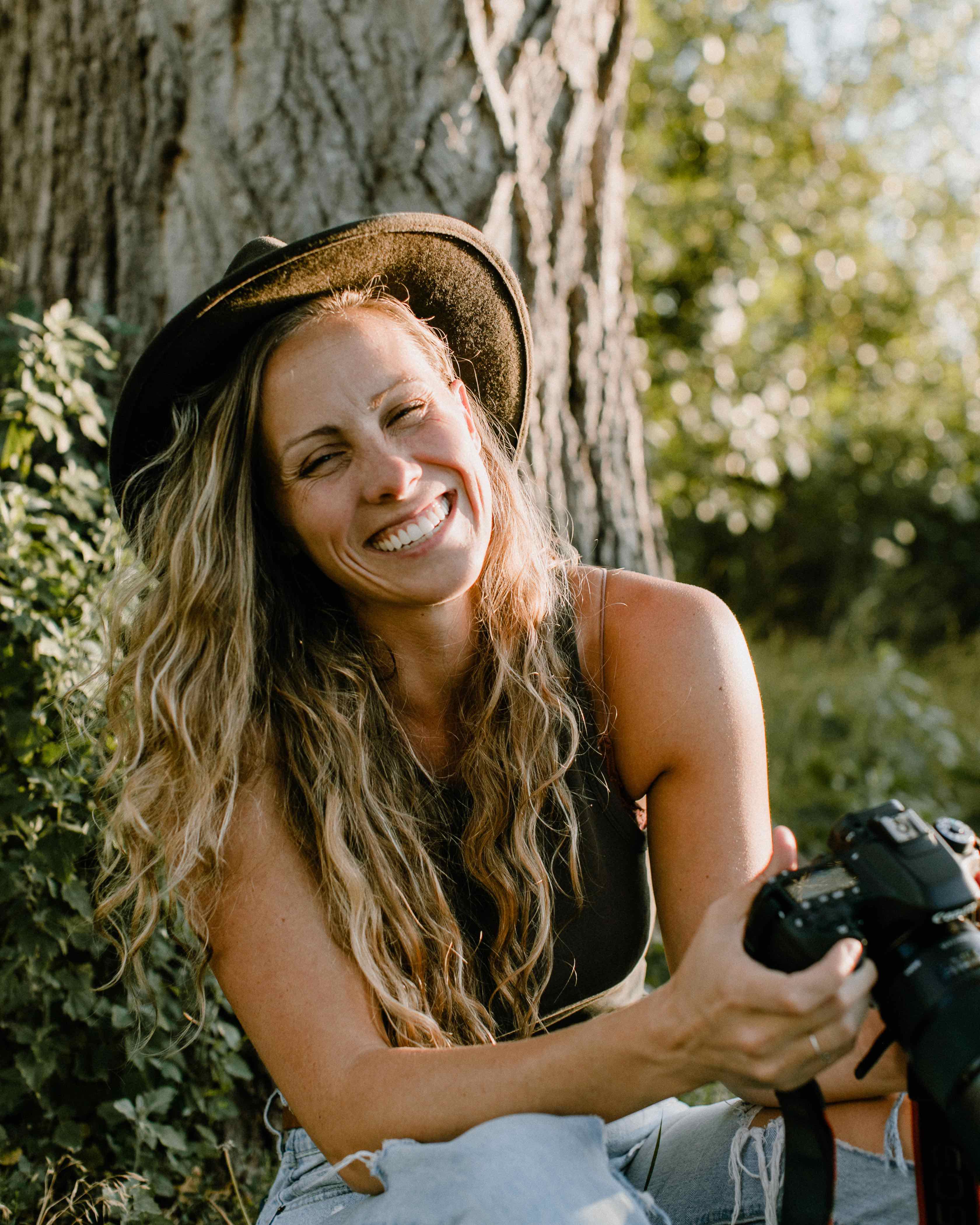 Woman is smiling while laughing and holding a camera. She is sitting down with her back against a tree and the setting sun is highlighting the side of her face and hair.