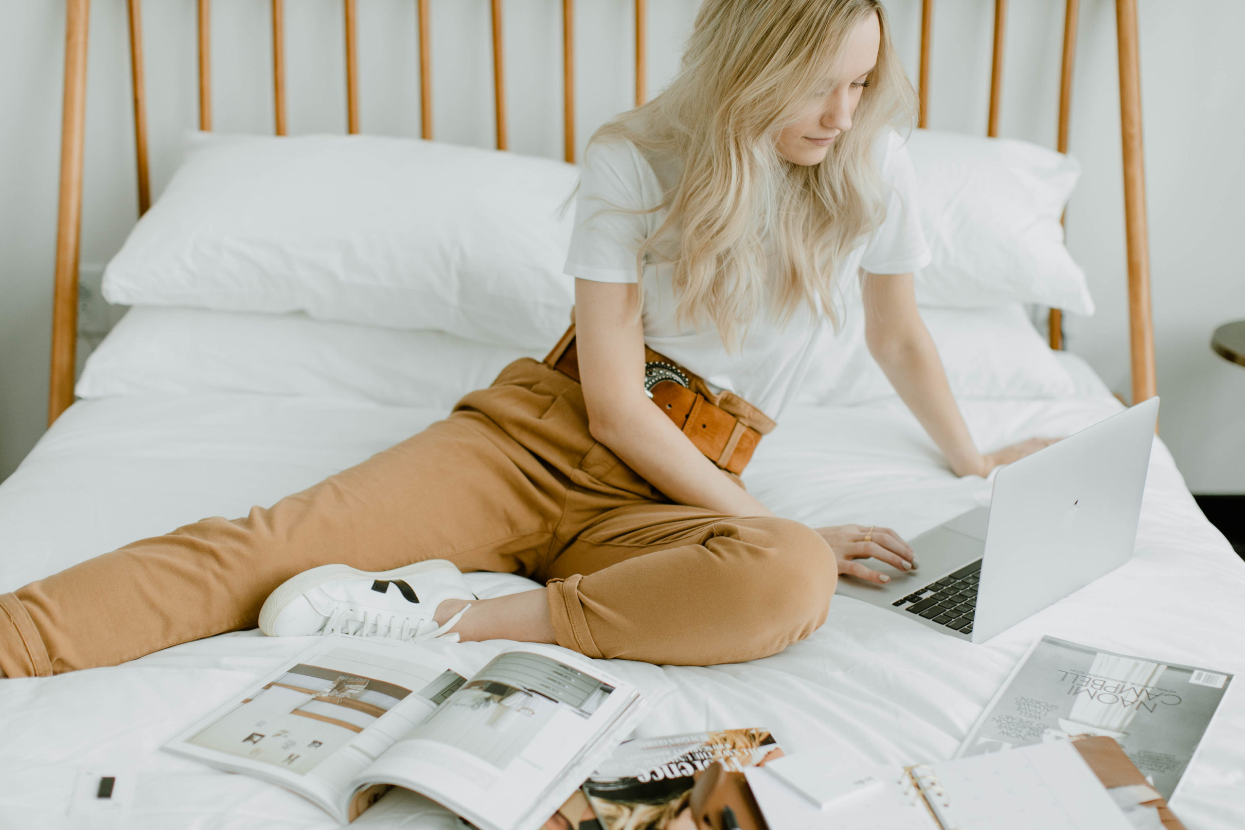 Image of a woman sitting on a bed surrounded by open magazines and working on a laptop during her brand photography session.